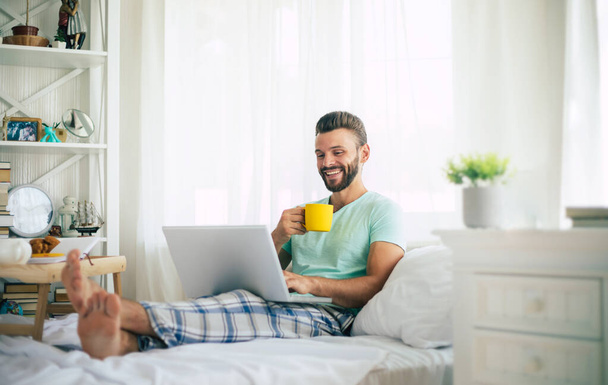 Handsome young bearded man in casual clothes is working at home while sitting on the bed. Confident guy with a laptop and smartphone drinks coffee in bedroom. - Foto, Imagem