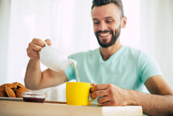 Happy young bearded man is sitting on his bed and has his breakfast with tea or coffee - 写真・画像