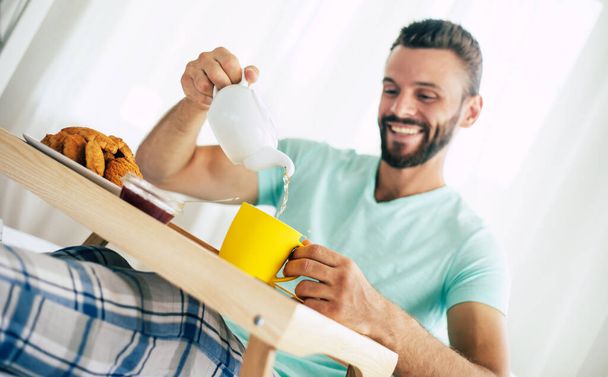 Happy young bearded man is sitting on his bed and has his breakfast with tea or coffee - Fotó, kép
