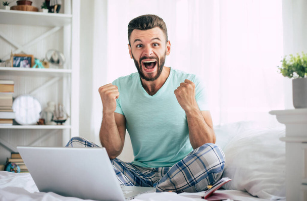 Excited happy win fan bearded man with a laptop in a big white bed at home is shouting while the celebration - Foto, Bild