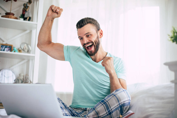 Excited happy win fan bearded man with a laptop in a big white bed at home is shouting while the celebration - Valokuva, kuva