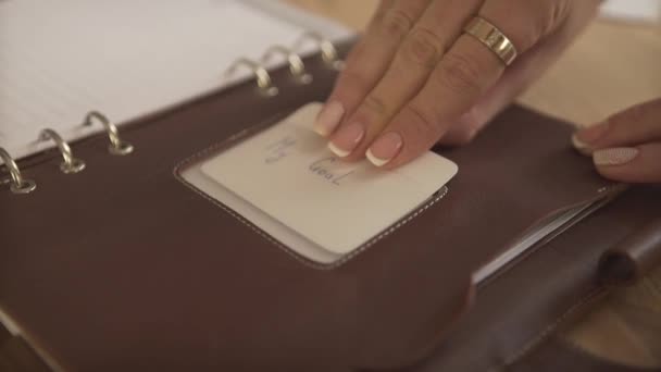 Close up shot of woman putting card sticker in brown leather case cover of organizer journal daily planner on wood table - Footage, Video