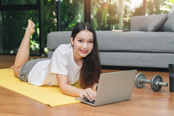 Gyönyörű fiatal nő néz fitness video tutorials online laptopon, miközben feküdjön le a földre a nappaliban. - Fotó, kép