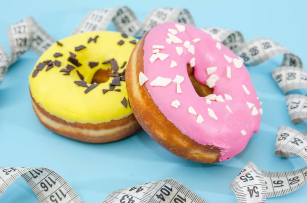 two donuts with yellow and pink frosting and measuring tape isolated on blue background. The concept of losing weight and the harm of sweets. Diet. - Photo, Image