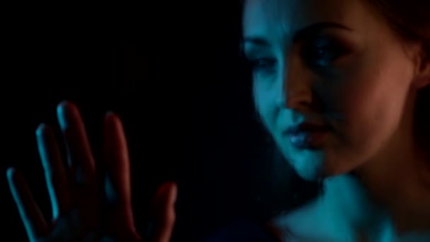 Dromerig aantrekkelijk Kaukasische jonge vrouw kijkt naar glas raam tekent een hart met vingertoppen in de nacht blauw neon licht - Video