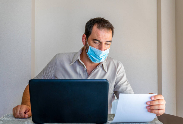 Sakallı, bej gömlekli ve maske takan bir adam kendini koronavirüsten korumak için oturma odasındaki bilgisayardan endişelenen faturalara bakıyor. Umutsuzluk konsepti - Fotoğraf, Görsel