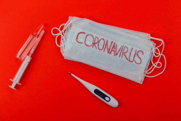 Máscara médica protetora com a inscrição coronavírus, seringas, ampolas e termômetro em um fundo vermelho. Máscara protetora como precaução para a propagação do coronavírus Covid-19 em todo o mundo. - Foto, Imagem