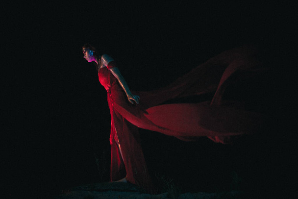 Cinematic goddess stands in the dark, her red dress developing on the wind - Fotoğraf, Görsel