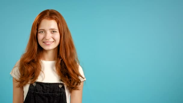 Young woman with red hairstyle in overall looking to camera. Copy space. Portrait of cute beautiful girl on blue wall studio background. - Séquence, vidéo