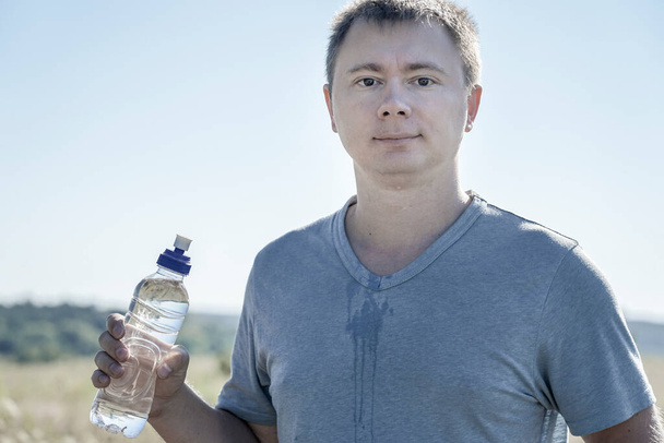 een man met een bril en een t-shirt houdt een plastic fles water in zijn handen - Foto, afbeelding
