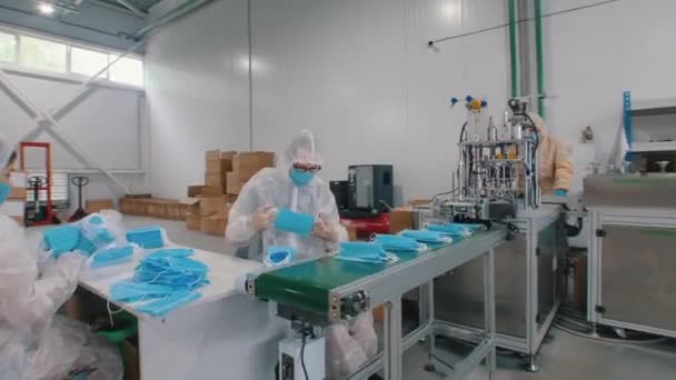 Producción industrial de máscaras médicas protectoras - los trabajadores en traje de protección y guantes empaca las máscaras juntos - Imágenes, Vídeo