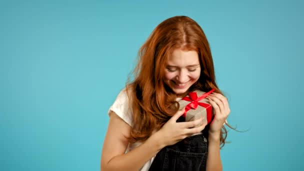 Cute young woman in overall holding gift box with bow, she wonders what is inside. Blue wall background. Girl smiling, she is happy to get present. - Video
