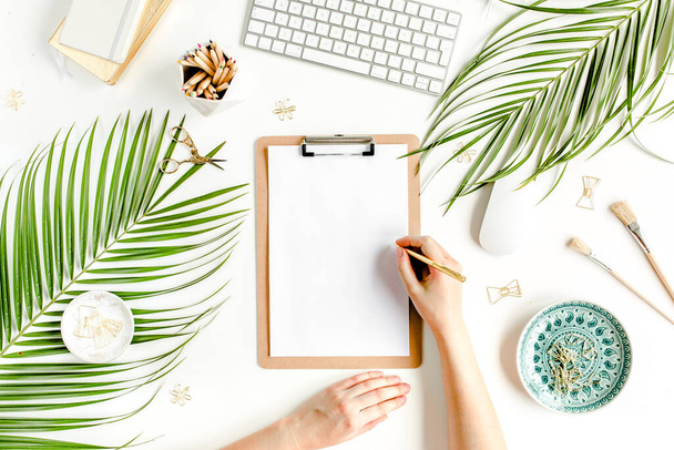 Female workspace with female hands, clipboard, tropical palm leaves, computer, accessories on white background. Stylized womens desk. Flat lay. Top  - 写真・画像