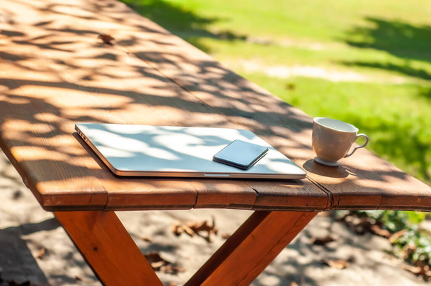 Vista su un computer portatile chiuso e una tazza di caffè smartphone sul tavolo in giardino in un ambiente domestico o scolastico in una giornata di sole. - Foto, immagini