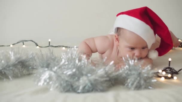 merry christmas christmas and happy new year, infants, childhood, holidays concept - close-up naked 6 month old newborn baby in santa claus hat on his tummy crawls with decorations on christmas tree. - Materiał filmowy, wideo