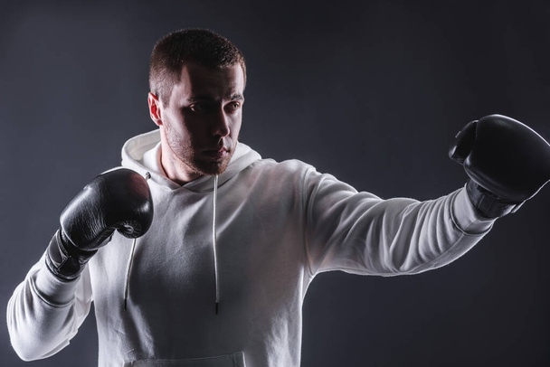 Young man in boxing gloves and a white sweatshirt on a dark background in the studio. The concept of victories and battles - Φωτογραφία, εικόνα
