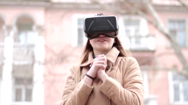 Młoda szczęśliwa dziewczyna w wirtualnej rzeczywistości okulary zestaw słuchawkowy zabawy na zewnątrz na ulicy w beżowym płaszczu - Materiał filmowy, wideo