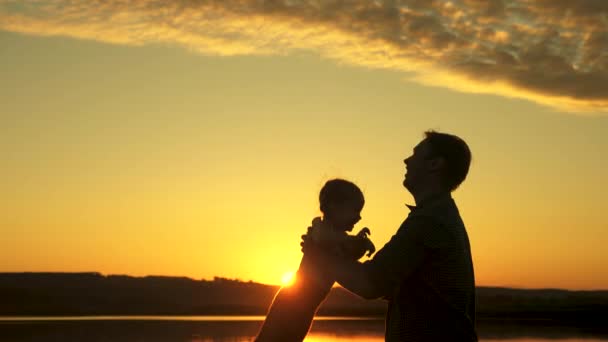 sziluett apa és egészséges gyermek repül a nap felett. Apa feldobja a boldog kislányát a partra, és szórakozik naplementekor. Apa vízen játszik a gyerekkel. boldog család és gyermekkor - Felvétel, videó