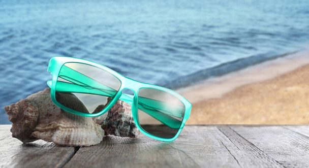 Shells and stylish sunglasses on wooden table near sea with sandy beach - Photo, image