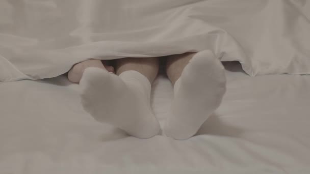 close-up shot van vrouwelijke blote voeten aanraken mannelijke voeten in witte sokken onder deken op bed met witte lakens - Video