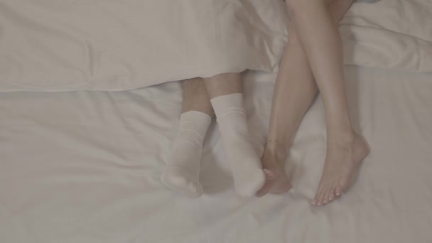 Männerbeine in weißen Socken von zwei barfüßigen Frauen berührt, die unter einer Decke auf weißen Bettlaken auf dem Bett liegen - Filmmaterial, Video