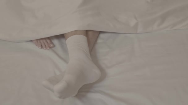 man benen in wit sokken aangeraakt door twee vrouwen met blote voeten liggend op bed op wit bed lakens onder deken in close up - Video