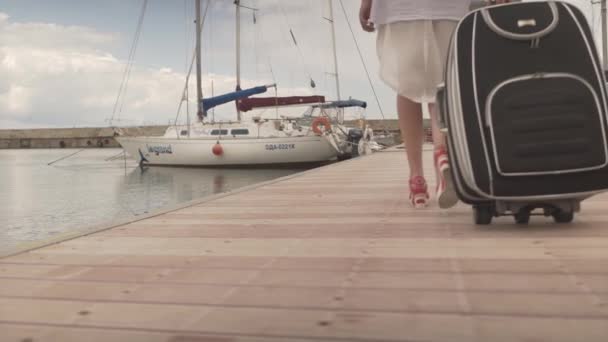 Молодая привлекательная женщина возвращается на высоких каблуках прогуливаясь по деревянному мосту с тяжелым багажом в гавани яхт - Кадры, видео