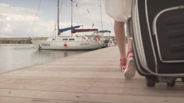 Молодая привлекательная женщина на высоких каблуках поворачивает назад с тяжелым багажом ходьба по деревянному мосту в гавани яхт - Кадры, видео