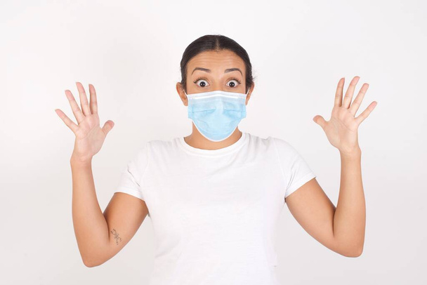 Optimistic  Young arab woman wearing medical mask standing over isolated white background raises palms from joy, happy to receive awesome present from someone, shouts loudly, Excited caucasian female screaming. - Foto, Bild