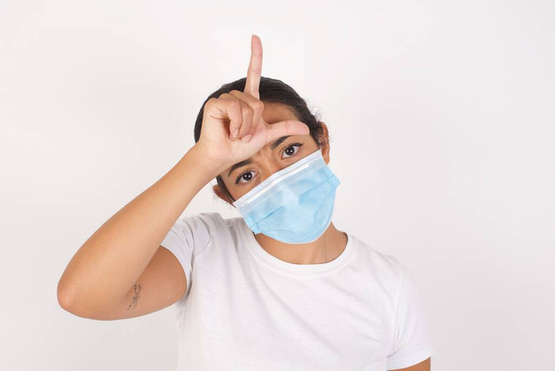 Mladá arabka žena nosí lékařskou masku stojící nad izolovaným bílým pozadím dělat si legraci z lidí s prsty na čele dělat ztroskotanec gesto posměch a urážlivé. - Fotografie, Obrázek