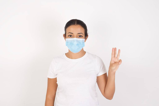 Mladá arabka žena nosí lékařskou masku stojící nad izolovaným bílým pozadím ukazující a ukazující prsty číslo tři a usmívající se sebevědomě a šťastně. - Fotografie, Obrázek