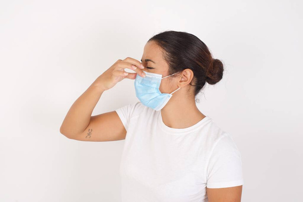 隔離された白い背景の上に立っている医療用マスクを身に着けている若いアラブの女性は、臭いと嫌な、耐え難い匂いを嗅ぎ、鼻の上に指で息を保持します。匂いが悪い - 写真・画像