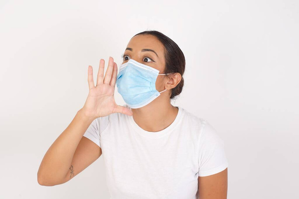 Young arab woman wearing medical mask standing over isolated white background shouting and screaming loud to side with hand on mouth. Communication concept. - Photo, Image