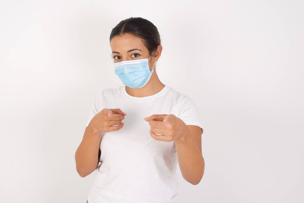 Junge arabische Frau mit medizinischer Maske, die vor isoliertem weißem Hintergrund steht und mit dem Finger in die Kamera zeigt, mit fröhlichem und lustigem Gesicht. Gute Energie und Stimmung.  - Foto, Bild