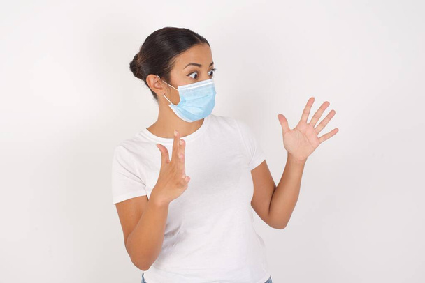 Mulher árabe jovem usando máscara médica em pé sobre fundo branco isolado gritos em voz alta, mantém os olhos abertos e as mãos tensas. - Foto, Imagem