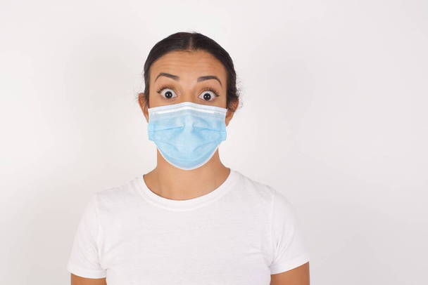 Oh my God. Surprised  Young arab woman wearing medical mask standing over isolated white background stares at camera with shocked expression exclaims with unexpectedness, - Photo, Image