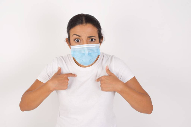 Photo of embarrassed Young arab woman wearing medical mask standing over isolated white background indicates at herself with puzzled expression, being shocked to be chosen to participate in competition, hesitates about something  - Foto, Imagem
