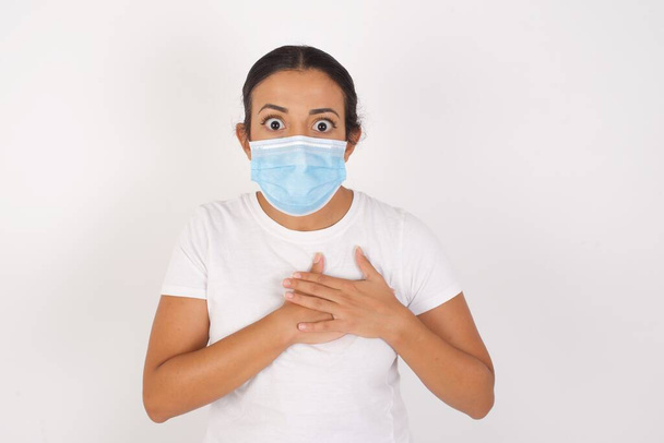 Scared Young arab woman wearing medical mask standing over isolated white background looks with frightened expression, keeps hands on chest, being puzzled to notice something strange,  People, hush reaction and emotions. - Foto, Bild