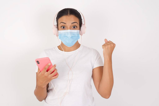 Pozitivní Mladá arabská žena v lékařské masce stojící nad izolovaným bílým pozadím drží moderní mobilní telefon připojen k sluchátkům, sevře pěst z dobrých emocí, zvolá radostně,  - Fotografie, Obrázek