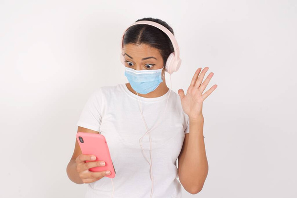 Mladý arab žena na sobě lékařská maska stojící nad izolované bílé pozadí držení v ruce buňky čtení prohlížení zpráv  - Fotografie, Obrázek