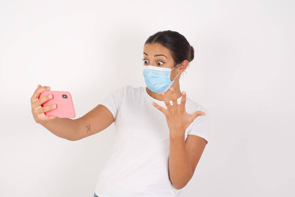 Páni! vzrušený mladý arab žena na sobě lékařská maska stojící nad izolované bílé pozadí ukazující mobilní telefon s otevřenou rukou gesto  - Fotografie, Obrázek