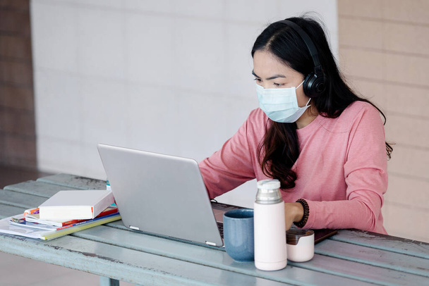 Młoda kobieta z maską na twarzy pracująca na laptopie w domu podczas pandemii Coronavirus lub COVID-19.. Uczeń skarbca studiuje online w domu. Zdjęcie z magazynu - Zdjęcie, obraz