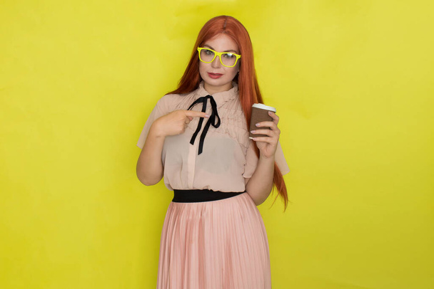 Red haired woman with glasses and office clothing holding cup of coffee or tea over yellow background, studio, style - Foto, afbeelding