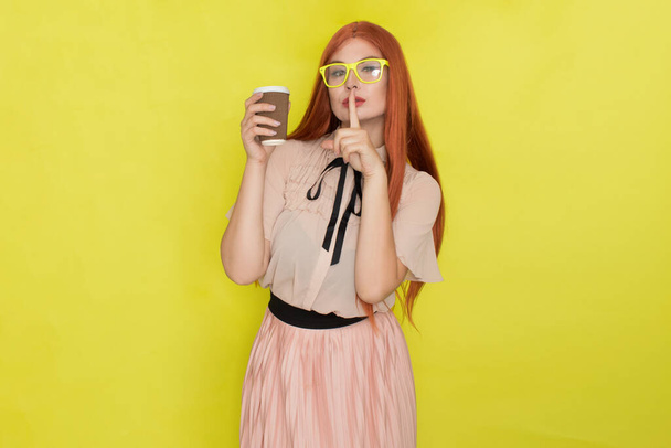 Red haired woman with glasses and office clothing holding cup of coffee or tea over yellow background, studio, style - Fotó, kép