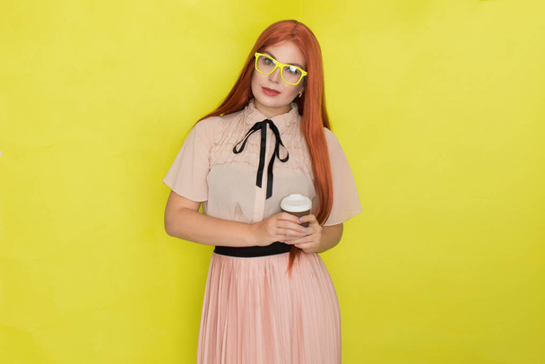 Red haired woman with glasses and office clothing holding cup of coffee or tea over yellow background, studio, style - Zdjęcie, obraz