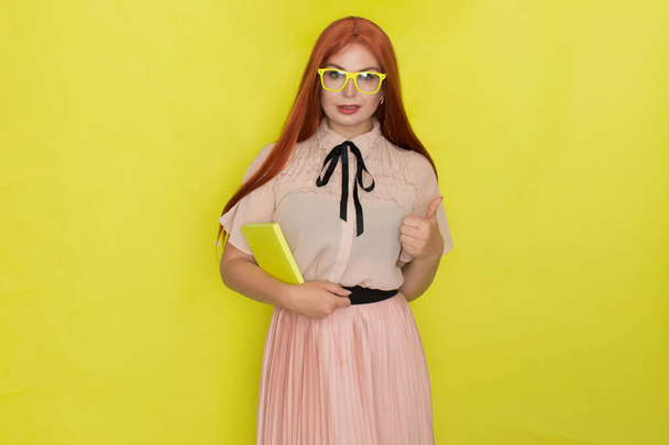 Red haired woman over yellow background holding book in her hands, studio shot, model posing - Foto, immagini