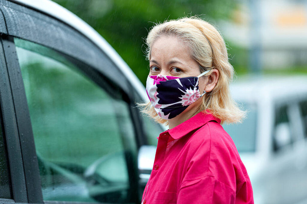 Pandemia COVID19 - Ritratto di profilo di donna mista che indossa una maschera facciale in tessuto fatto in casa fai da te per la protezione contro il Coronavirus e altre malattie infettive. Accanto alla macchina in un giorno di pioggia. - Foto, immagini