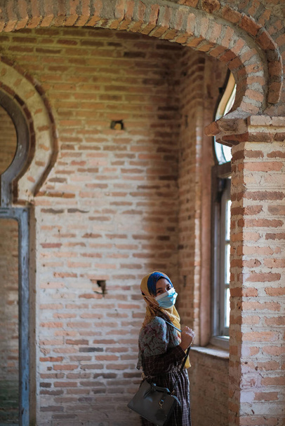 Πορτρέτο της νεαρής όμορφης Ασιάτισσας μουσουλμάνα με μαντίλα φορώντας ιατρική μάσκα στο παράθυρο - Φωτογραφία, εικόνα