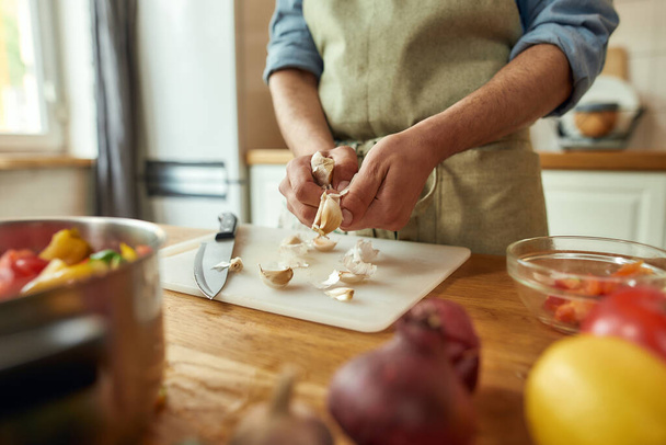 Close up of hands of man in apron peeling garlic while preparing healthy meal, soup in the kitchen. Cooking at home, Italian cuisine concept - Photo, image