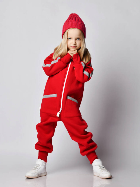 Full-growth portrait of blonde kid girl in red overall jumpsuit and winter hat swaying with her hands at her chin - Zdjęcie, obraz